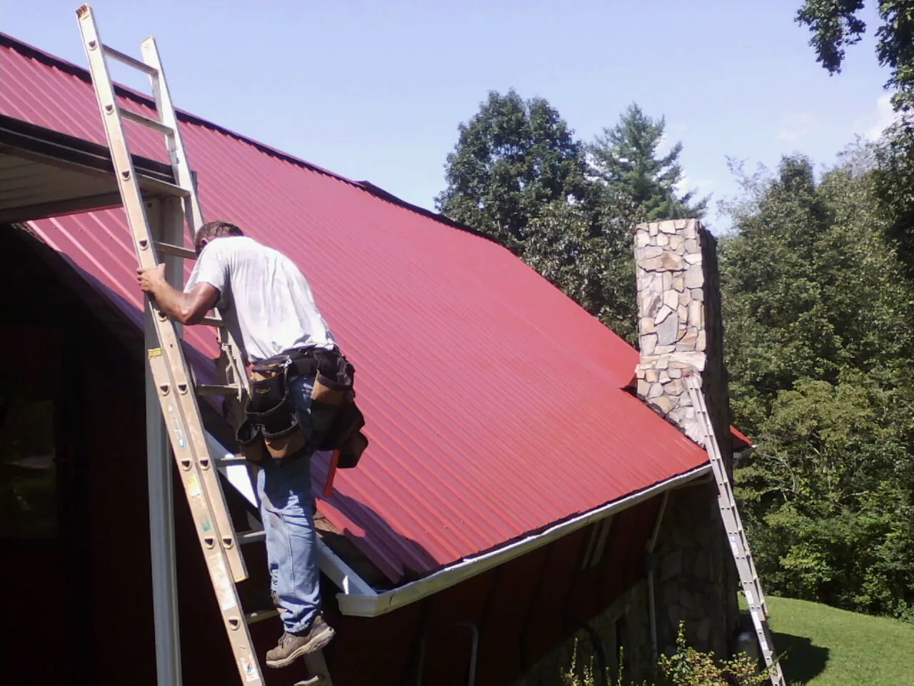 Roofer on a ladder beside a red metal roof and stone chimney — Soffit & Fascia Repair by Roofing Brothers in Voorheesville